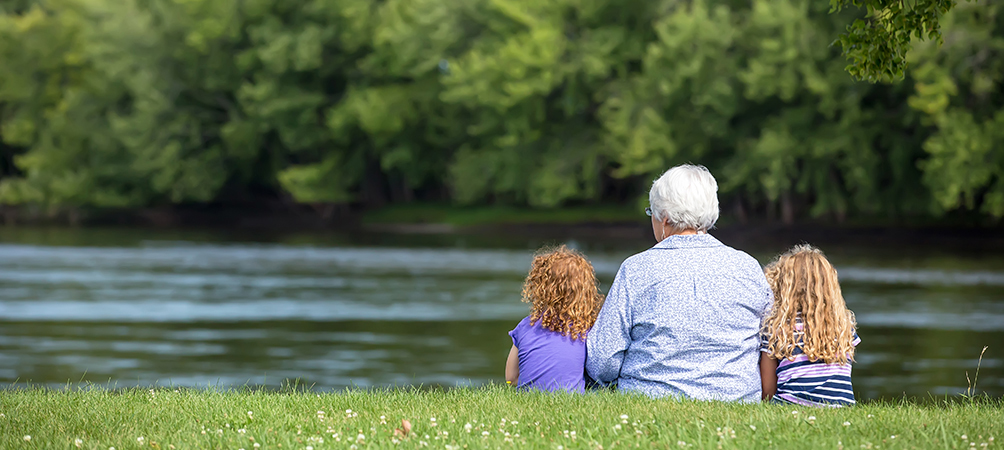 grandparent sitting in park in front of pond with grandchildren